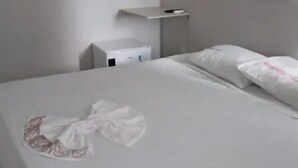 Una cama bien hecha con una colcha blanca y almohadas, una mesita de noche con una lámpara, y un estante pequeño sobre la cama.