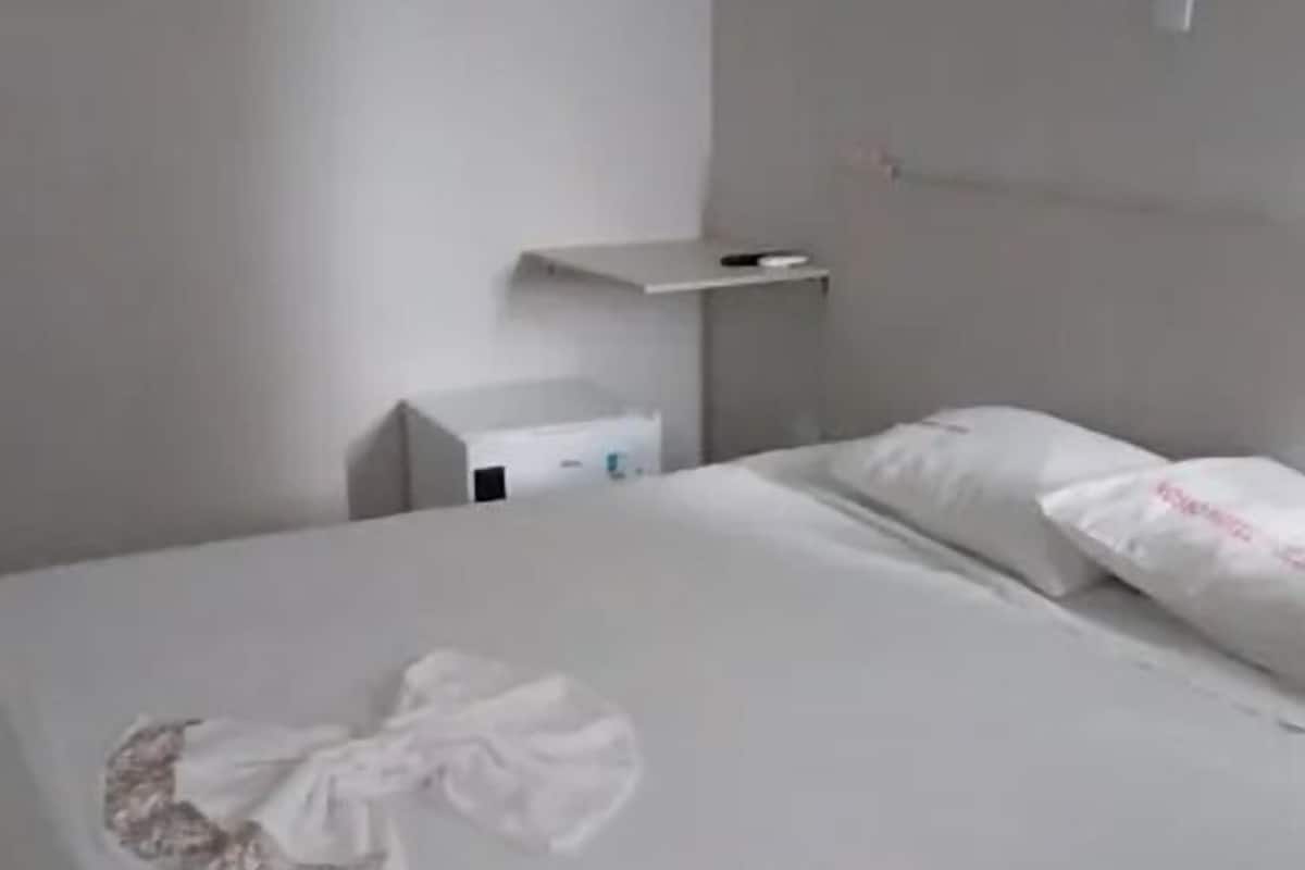 Uma cama bem arrumada com colcha branca e travesseiros, uma mesinha de cabeceira com abajur, e uma prateleira pequena acima da cama.