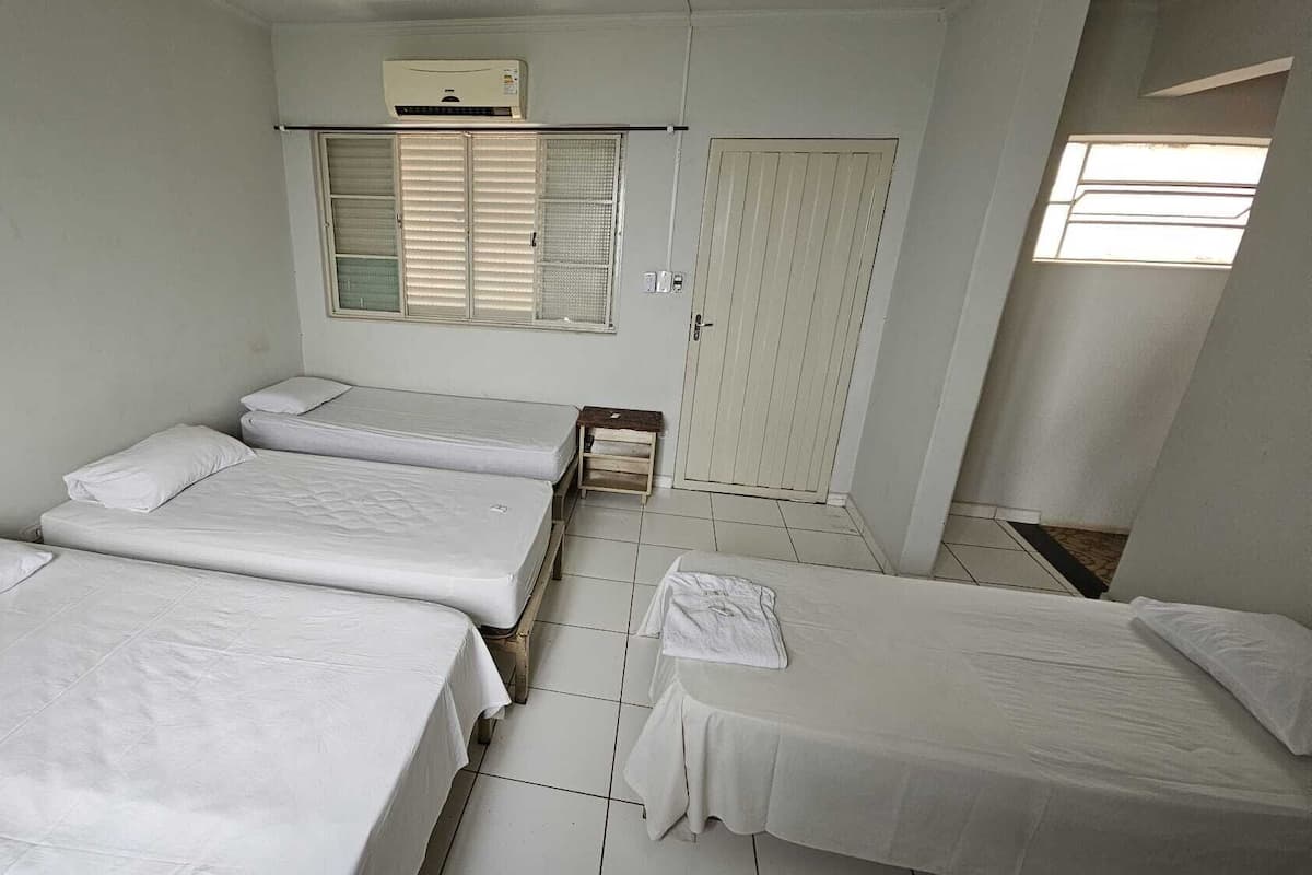 Quarto pequeno e limpo com três camas de solteiro, janela com persianas, porta e ar-condicionado fixado na parede.