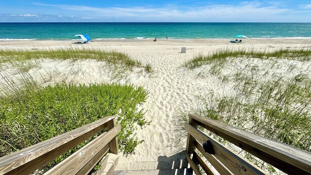 A beach with a wooden walkway leading to the water, clear skies, and a few umbrellas.
