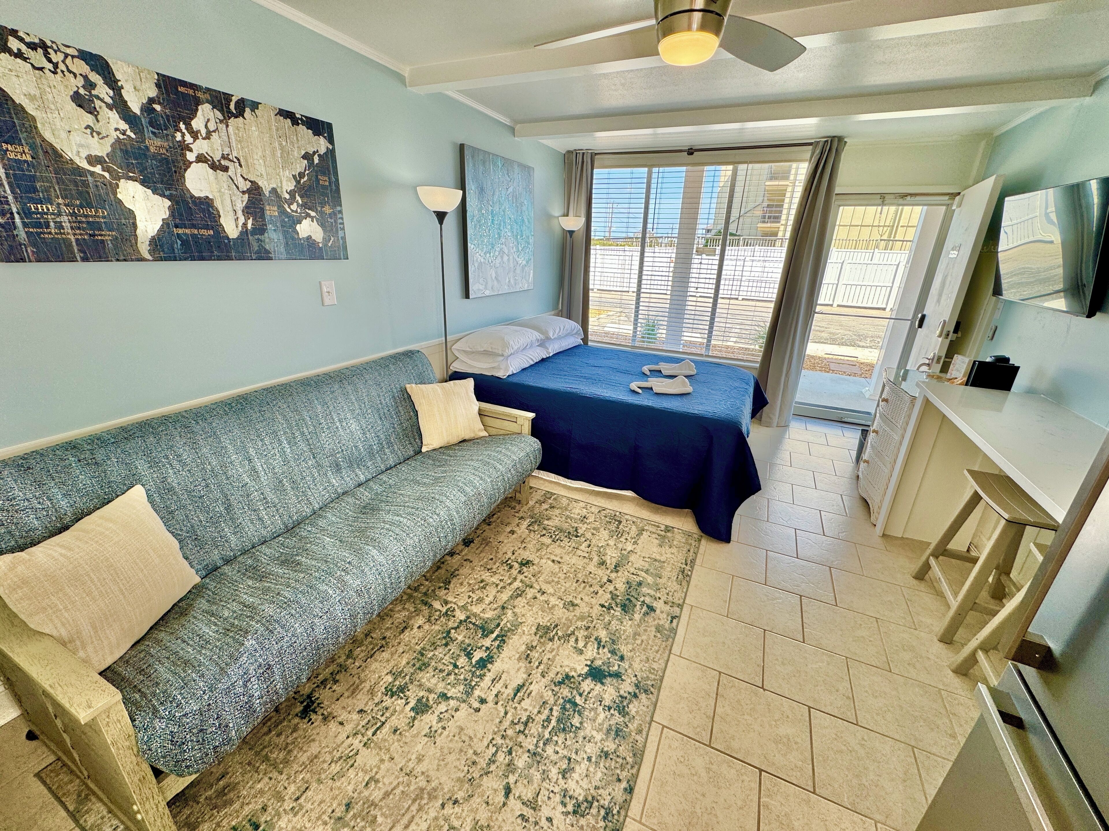 A room with a blue sofa, a bed with a blue bedspread, a world map on the wall, a floor lamp, and a view through a glass door.