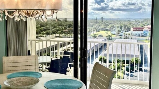 A dining area with a table set for two, a chandelier, and a view of a residential area with houses and greenery. - Paradise Sea's:  Updated 2B/2B with Ocean View (Cocoa Beach)