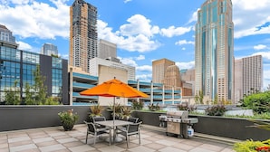 Um terraço no telhado com mesa, cadeiras, um guarda-sol laranja e uma churrasqueira.