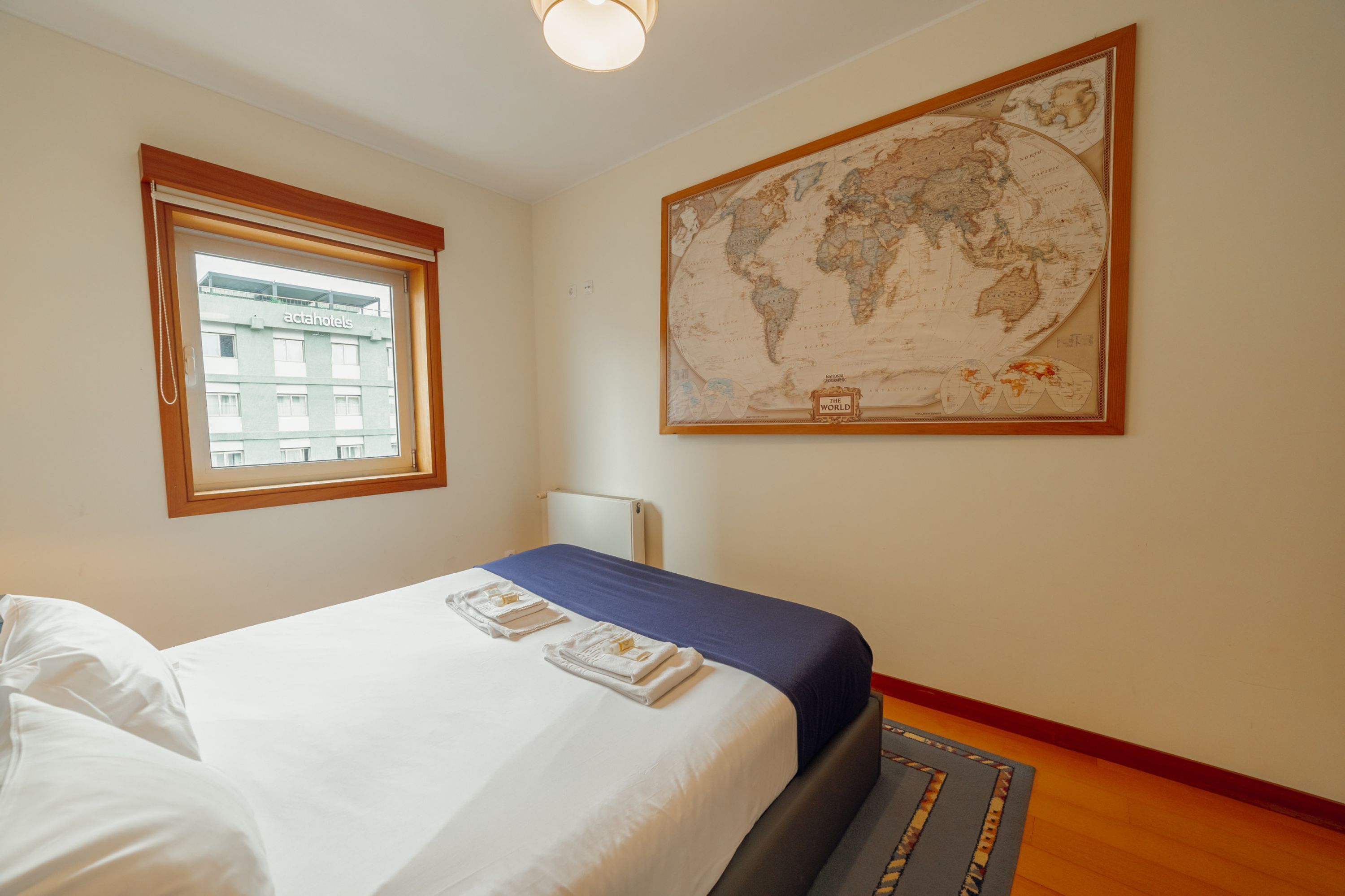 A hotel room with a bed, a window with a view of a building, and a large world map on the wall.