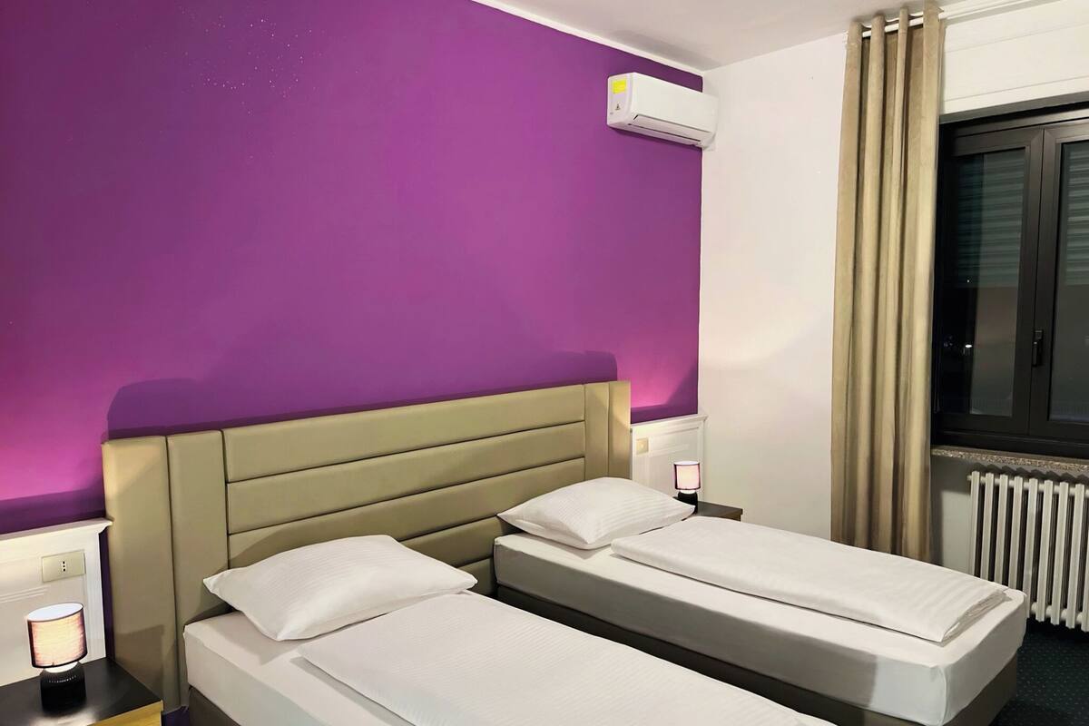 Habitación de hotel con dos camas individuales, cabecera, unidad de aire acondicionado, mesita de noche con lámpara y ventana con cortinas.
