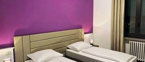 Habitación de hotel con dos camas individuales, cabecera, unidad de aire acondicionado, mesita de noche con lámpara y ventana con cortinas.