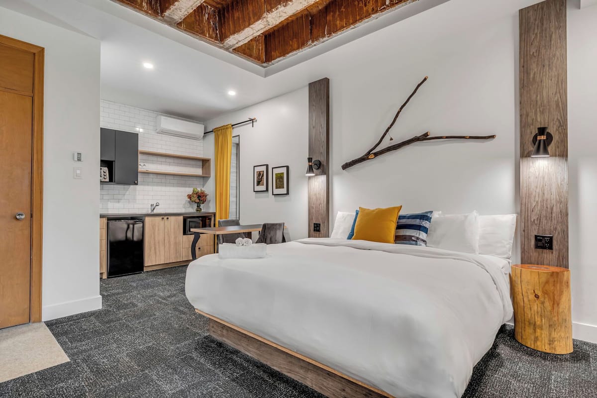 A modern hotel room with a large bed, a wooden headboard, a small wooden stool, a wall-mounted light, and a kitchen area in the background.
