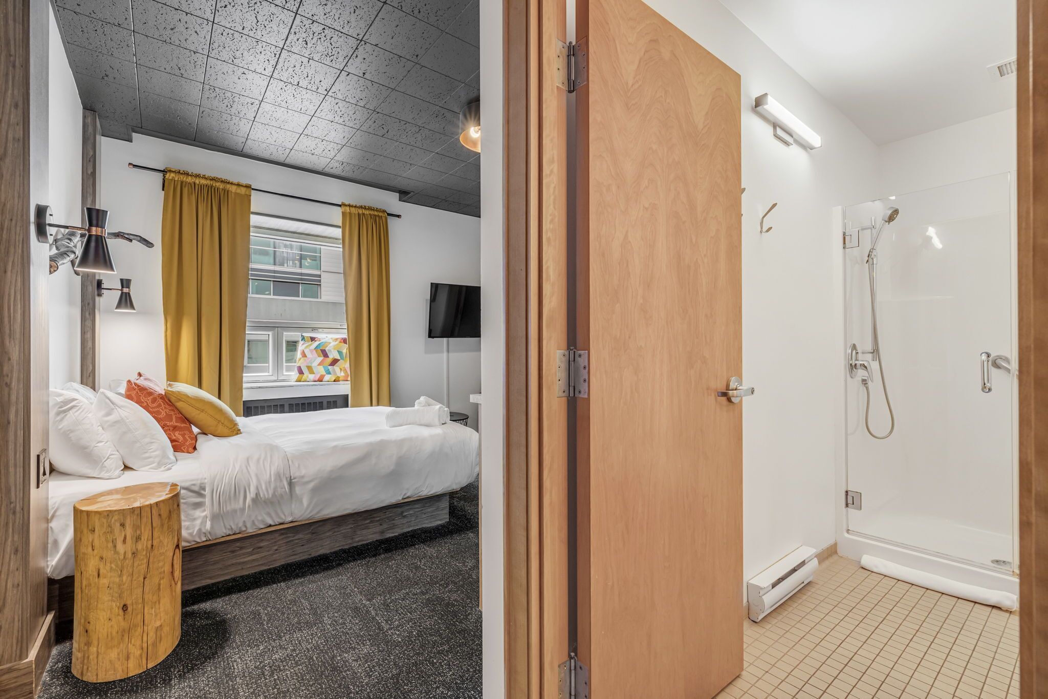 A hotel room with a bed, bedside table, and a wooden stool. There is a bathroom with a shower and a toilet.