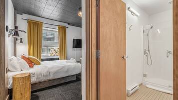 A hotel room with a bed, bedside table, and a wooden stool. There is a bathroom with a shower and a toilet.