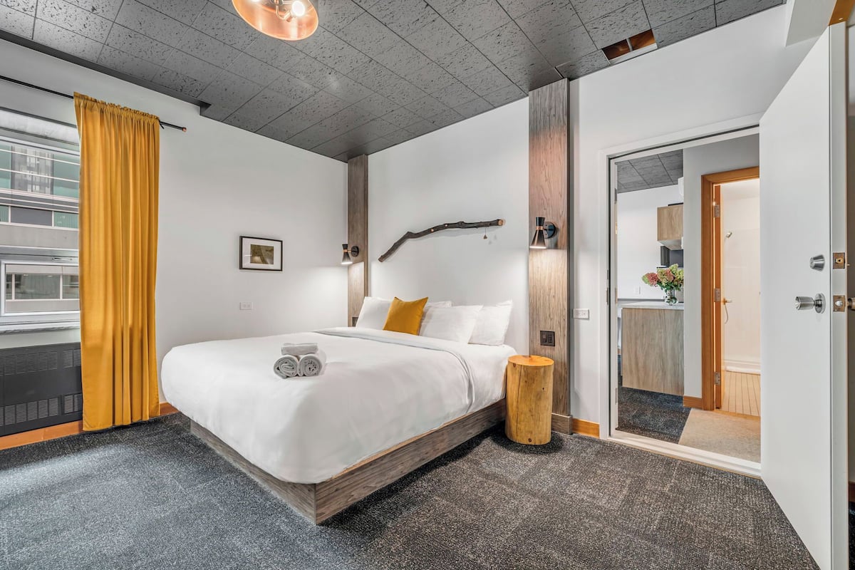 A modern bedroom with a large bed, a wooden bedside table, a wall-mounted light fixture, and a view of the outside through a window with yellow curtains.