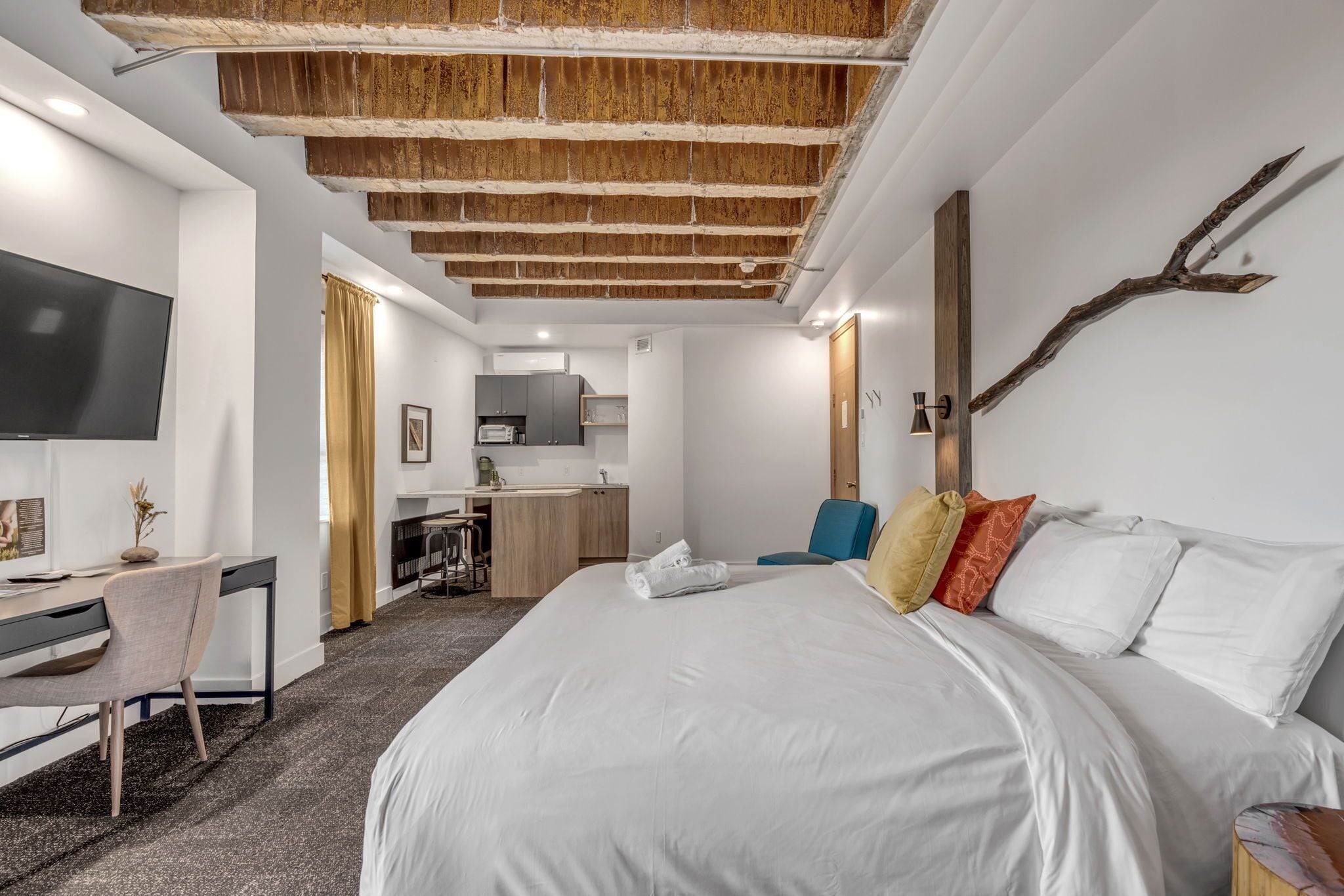 Habitación de hotel moderna con una cama grande, un escritorio, un televisor y una kitchenette al fondo.