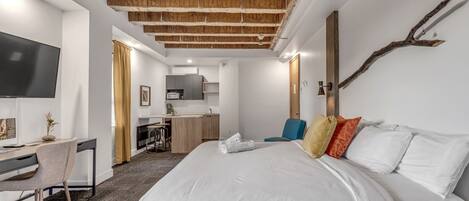 Habitación de hotel moderna con una cama grande, un escritorio, un televisor y una kitchenette al fondo.