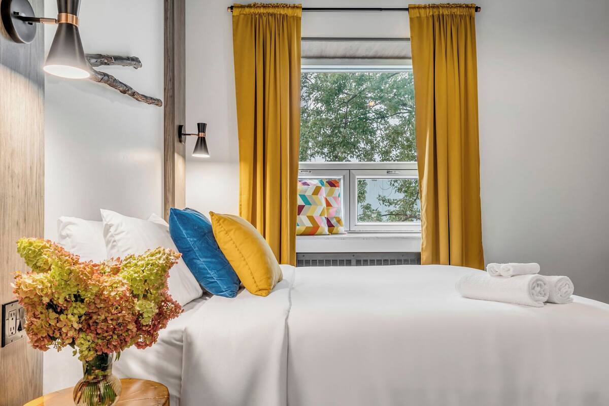A neatly made bed with white linens, a vase of flowers, and a window with yellow curtains.