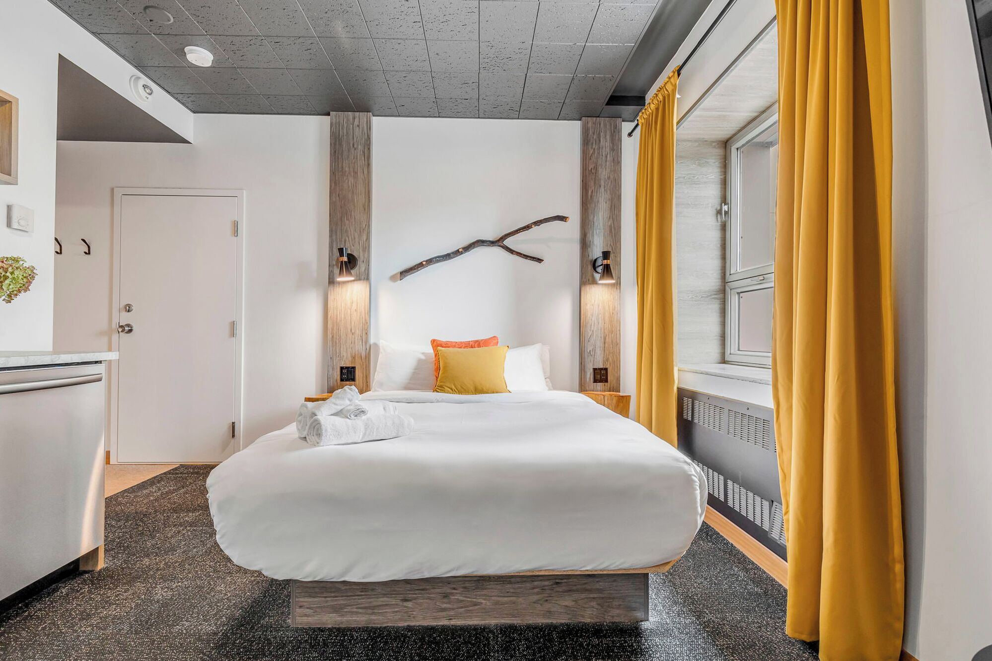 Une chambre d’hôtel avec un grand lit, une tête de lit en bois, deux lampes fixées au mur, une fenêtre avec des rideaux jaunes et une petite plante sur le mur de gauche.