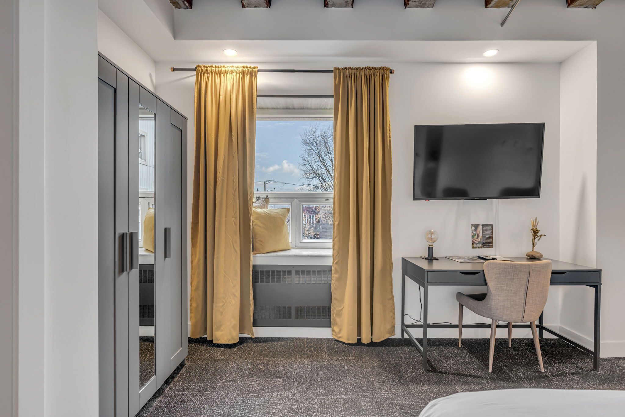A modern hotel room with a bed, a desk with a chair, a TV, and a window with curtains.