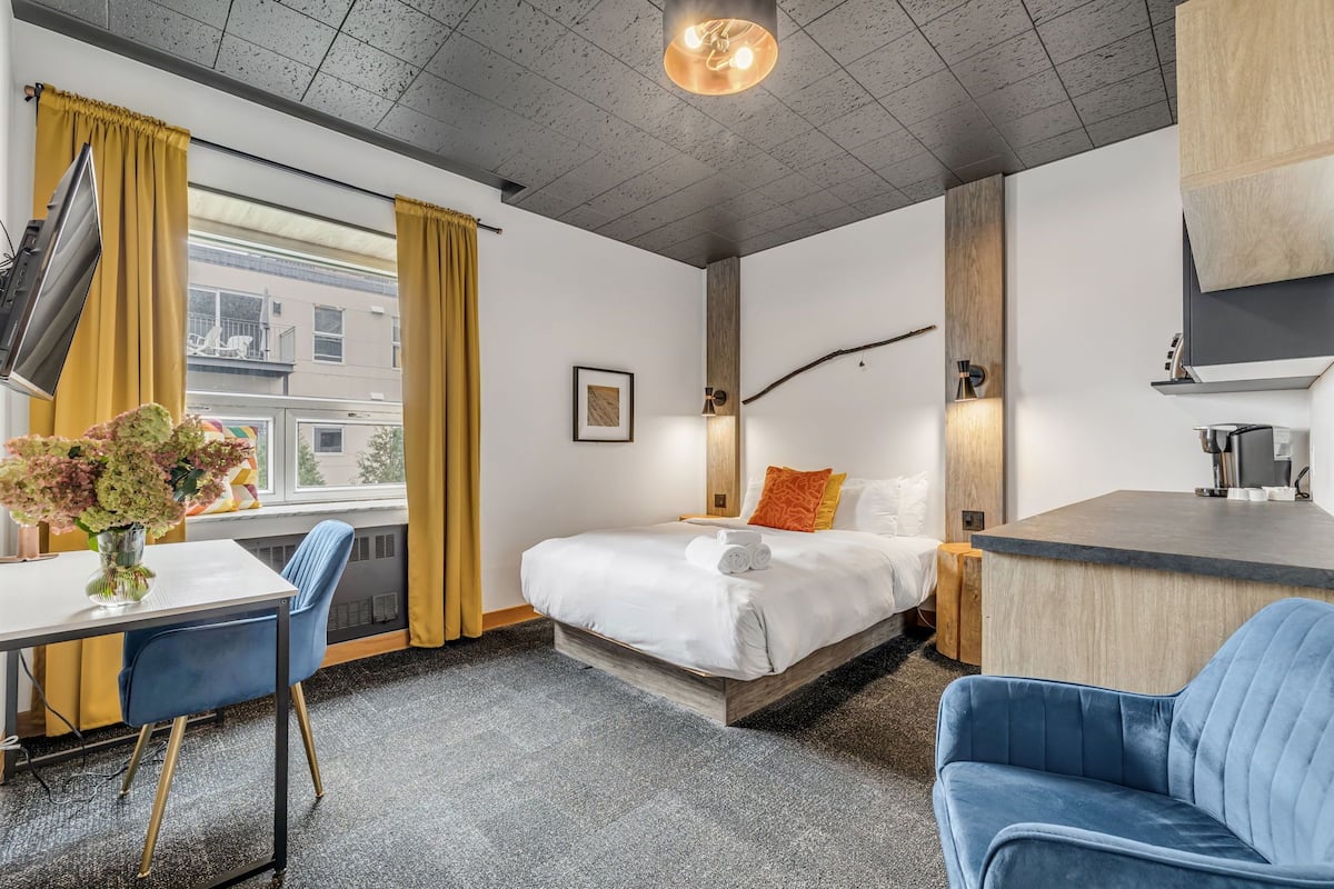 A modern hotel room with a bed, a desk, a chair, and a small kitchenette.
