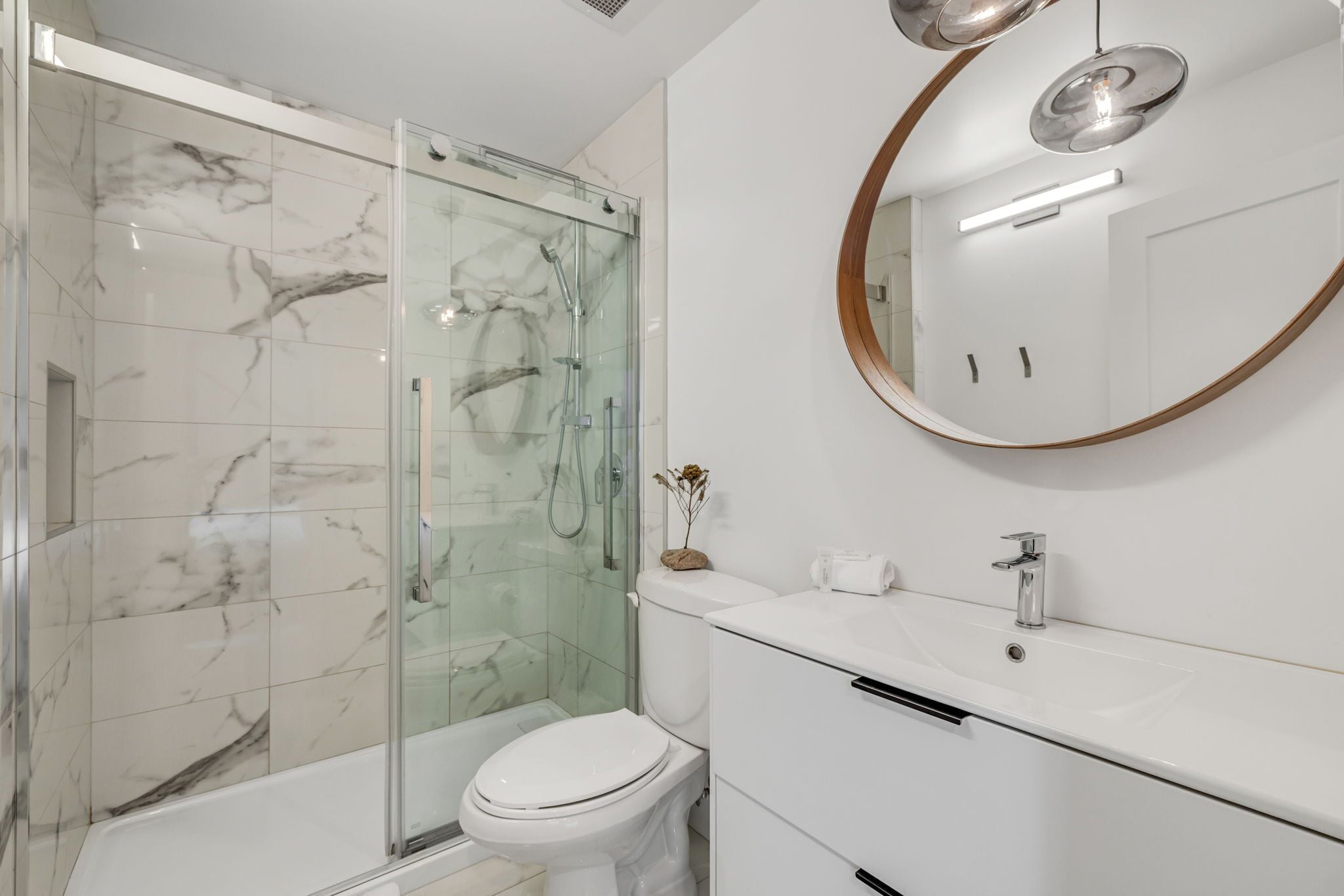 Un baño moderno con ducha de vidrio, inodoro blanco y lavamanos blanco con un espejo redondo encima.