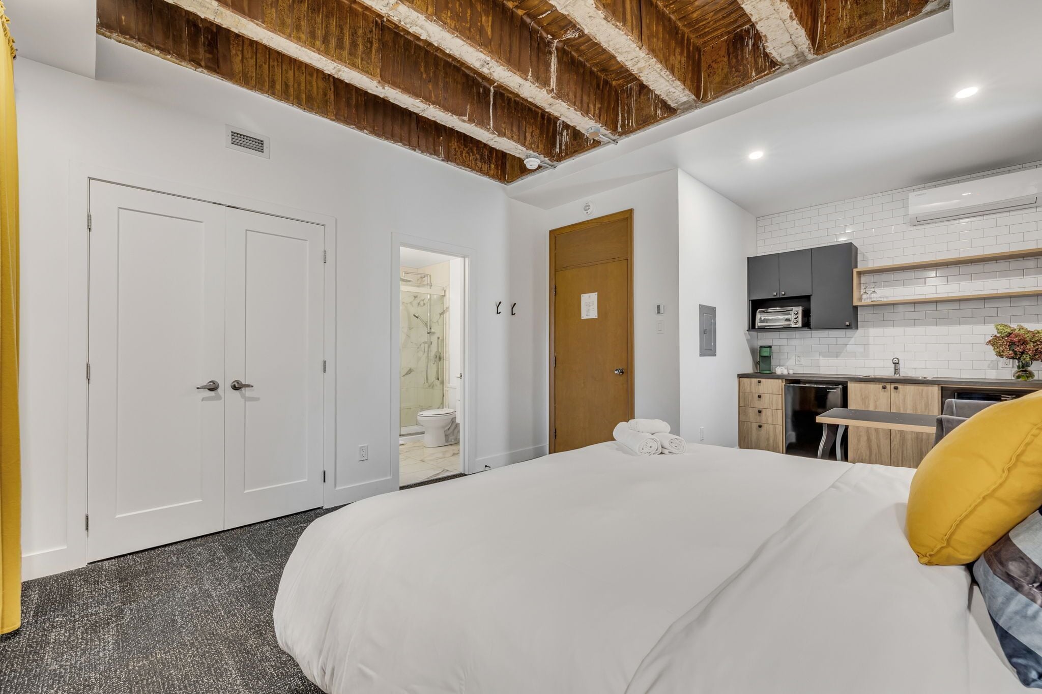 A modern bedroom with a large bed, a wooden ceiling, a small kitchenette, and a bathroom visible through an open door.
