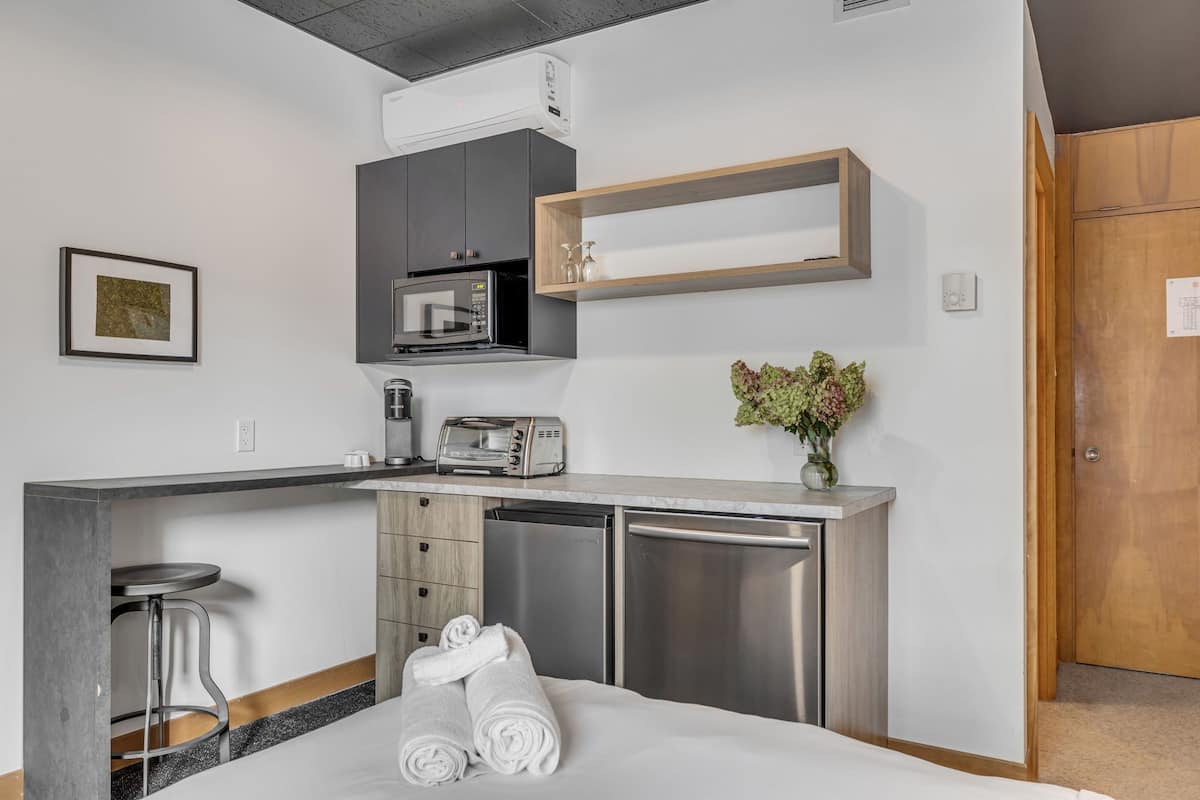 A compact kitchenette with a microwave, toaster, and a small refrigerator. There is a wooden shelf above the microwave and a small table with a vase of flowers.