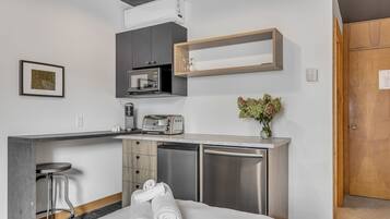 A compact kitchenette with a microwave, toaster, and a small refrigerator. There is a wooden shelf above the microwave and a small table with a vase of flowers.