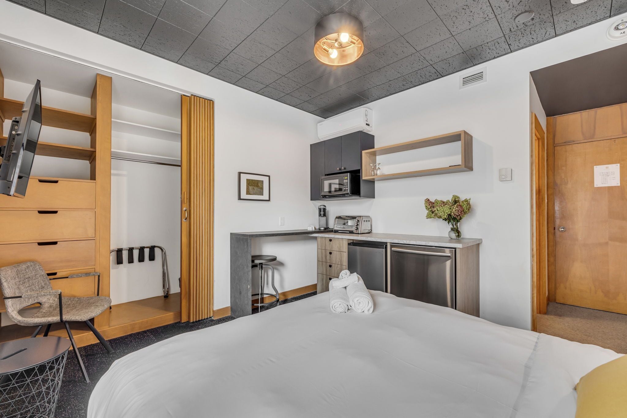 Una habitación de hotel moderna con una cama, un escritorio con microondas, una silla y una pequeña kitchenette.