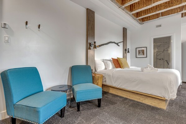 Ett modernt hotellrum med en säng, två blå fåtöljer, ett litet runt bord, ett träbord som används som sängbord och en vägglampa.
