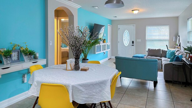 A dining area with a round table, yellow chairs, and a vase of flowers. There is a blue couch and a TV in the background.