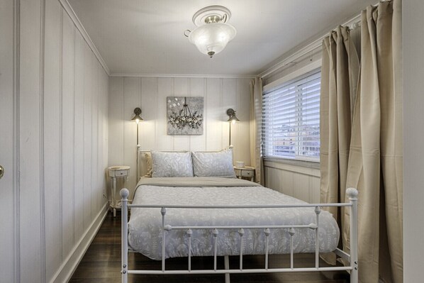A bedroom with a bed, bedside lamps, a chandelier, a window with curtains, and a wall-mounted artwork.