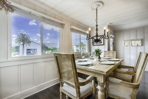 A dining room with a large table set for six, surrounded by wicker chairs, a chandelier, and large windows with a view of palm trees and buildings.