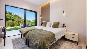 A modern bedroom with a large bed, a desk, and a view of greenery through a sliding door. - Villa Optima by Villa Plus (Ionian Islands)