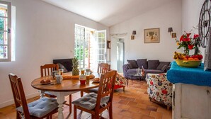 Un comedor acogedor con una mesa redonda de madera, sillas y un centro de mesa con piña. Se observa un sofá y un cuenco decorativo sobre una mesa al fondo.