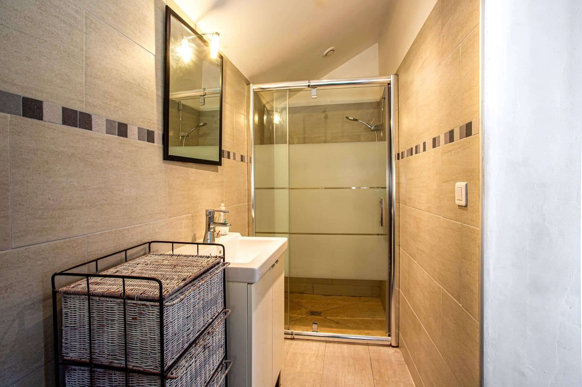 Une salle de bain moderne avec une douche à parois de verre, un lavabo avec un miroir au-dessus et un panier en osier posé sur un étagère en métal.