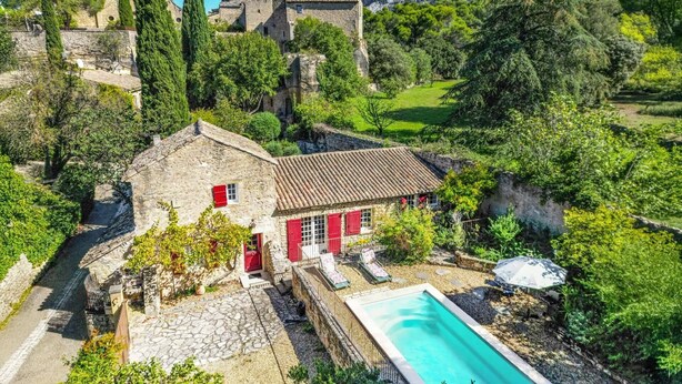 Casa de piedra con contraventanas rojas, piscina y montañas al fondo.