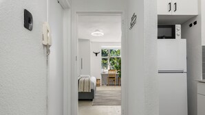 Un pasillo estrecho con una puerta blanca que da a un dormitorio con cama y escritorio.