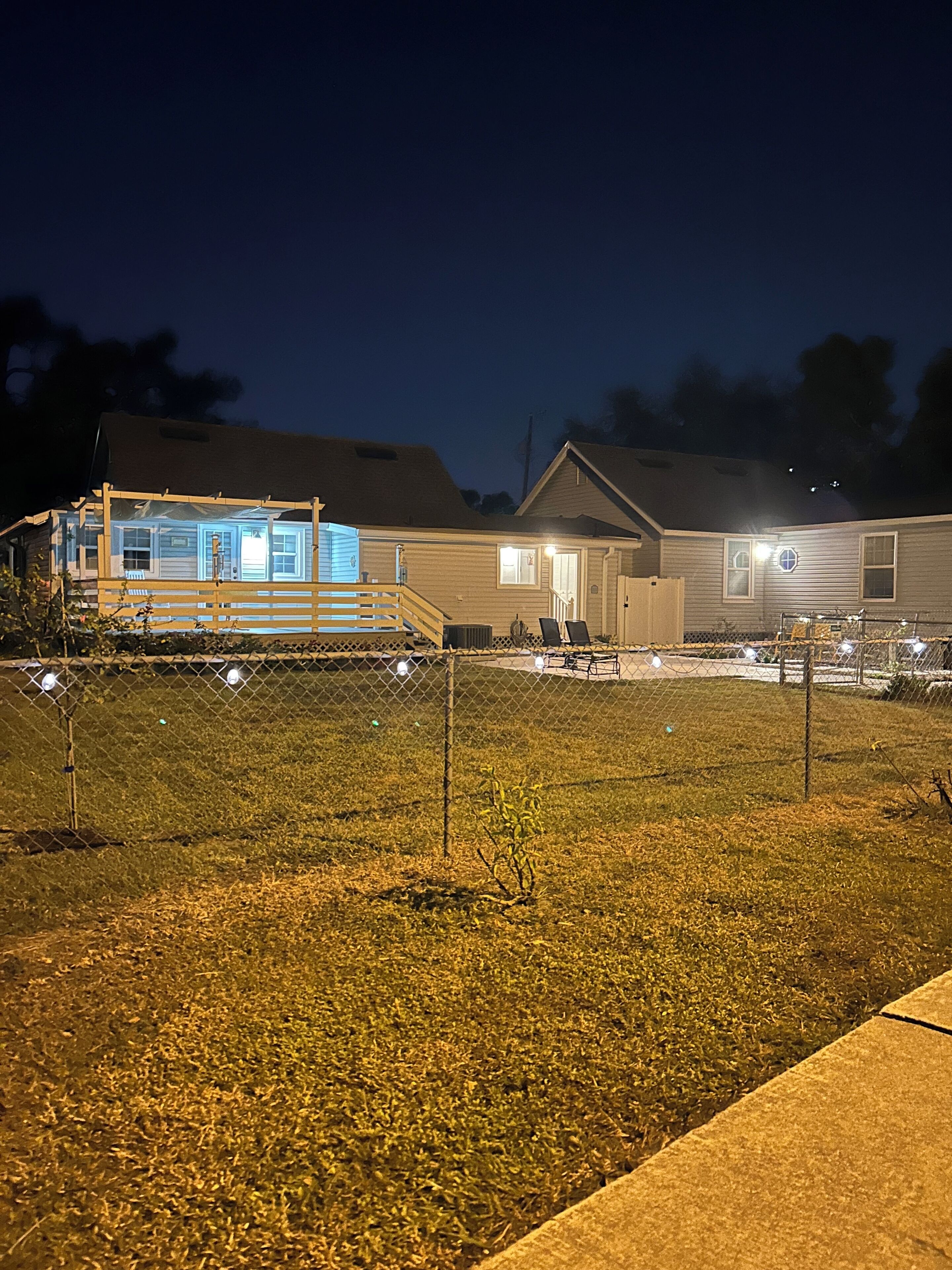 A fenced outdoor area with a building featuring a covered porch and a separate structure with a blue-lit entrance.
