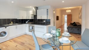 A modern kitchen with white cabinets, a built-in oven, and a washing machine. There is a dining area with a glass table and blue chairs. - Cosy flat in Leigh - Pass the Keys (Southend-on-Sea)