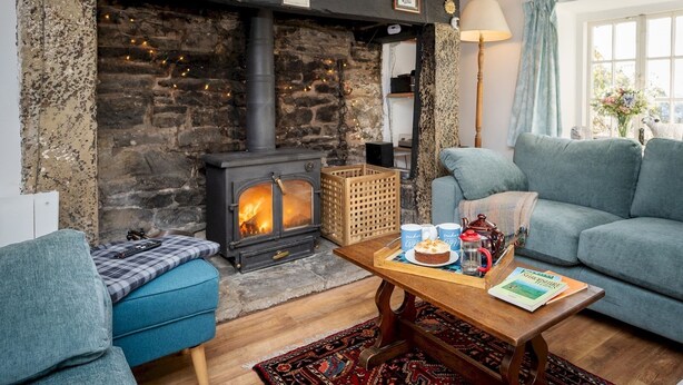 Un salon chaleureux avec une cheminée en pierre, un canapé bleu, une table basse en bois sur laquelle sont posés des livres et une théière, et un tapis à motifs.