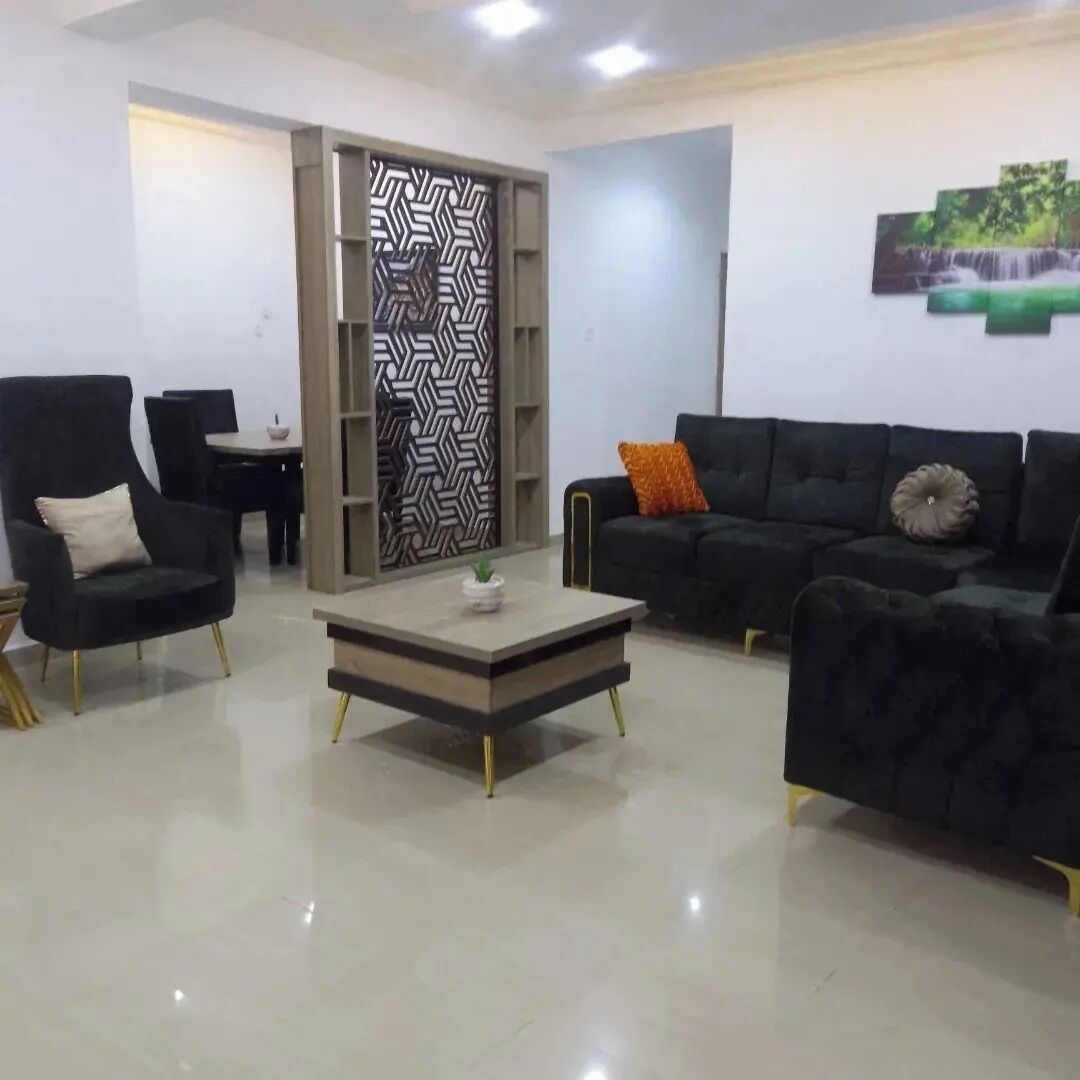 A modern living room with a black armchair, a black sofa with orange and beige cushions, a glass coffee table, and a patterned partition wall.