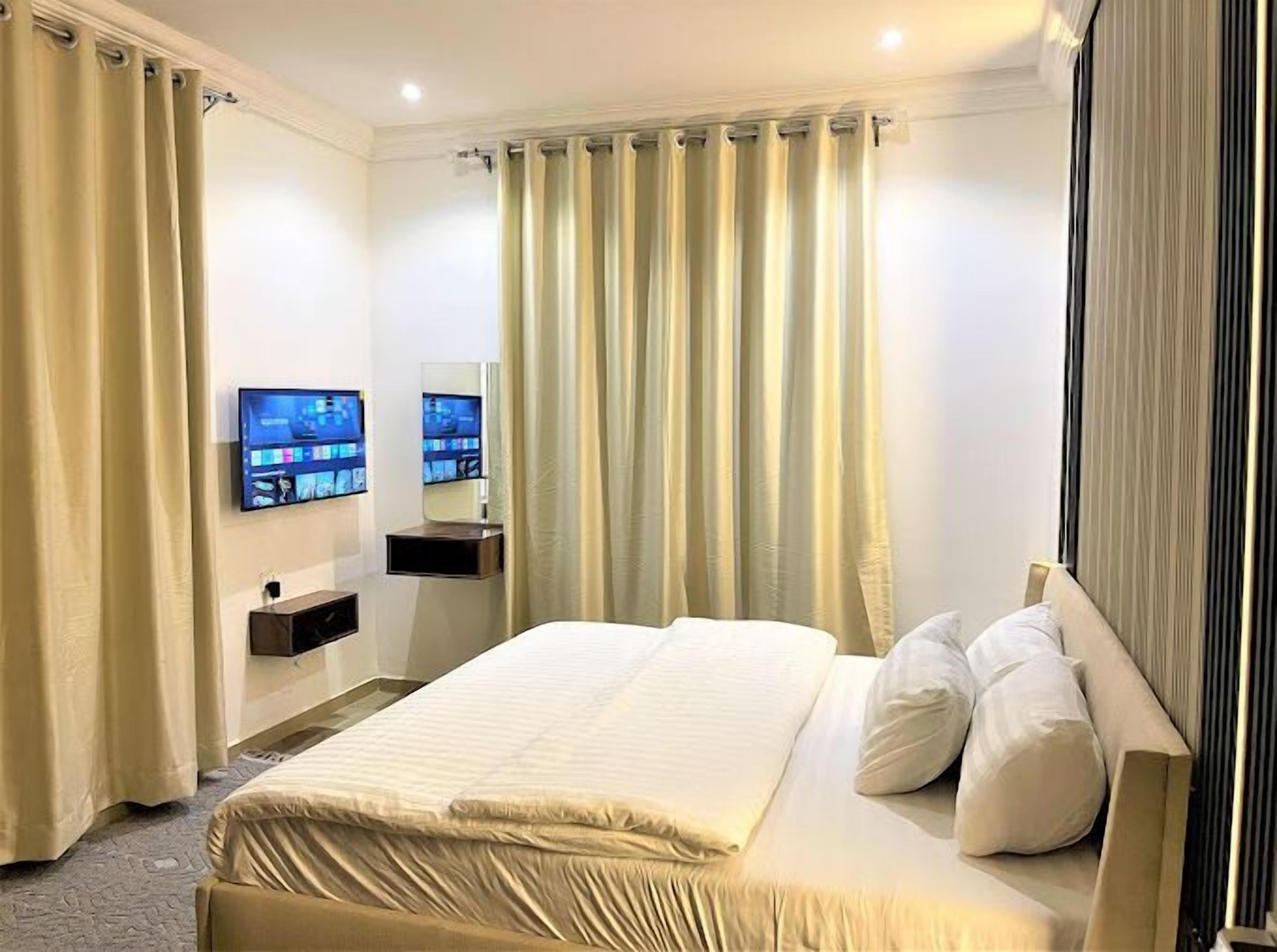 A hotel room with a bed, two wall-mounted TVs, a bedside table, and a large window with curtains.
