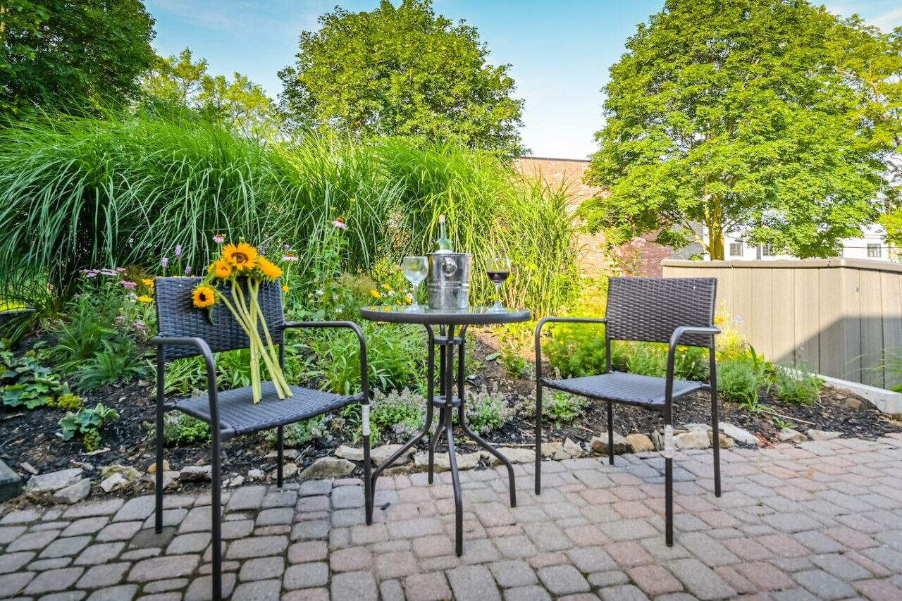An outdoor patio with two metal chairs and a small round table, surrounded by lush greenery and a brick pathway.