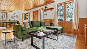 Interior acogedor de una cabaña con un sofá verde, techo de madera y un ventanal con vistas a árboles cubiertos de nieve.