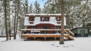 Una cabaña de dos pisos con terraza de madera y puerta verde, rodeada de árboles nevados.