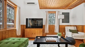 Una sala de estar acogedora con un sofá verde, una mesa de centro negra y un sillón blanco. Un televisor de pantalla plana está montado en la pared y hay una ventana con cortinas.