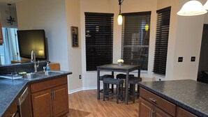 Una cocina con armarios de madera, encimera de granito, una mesa de comedor con sillas y un televisor negro montado en la pared.