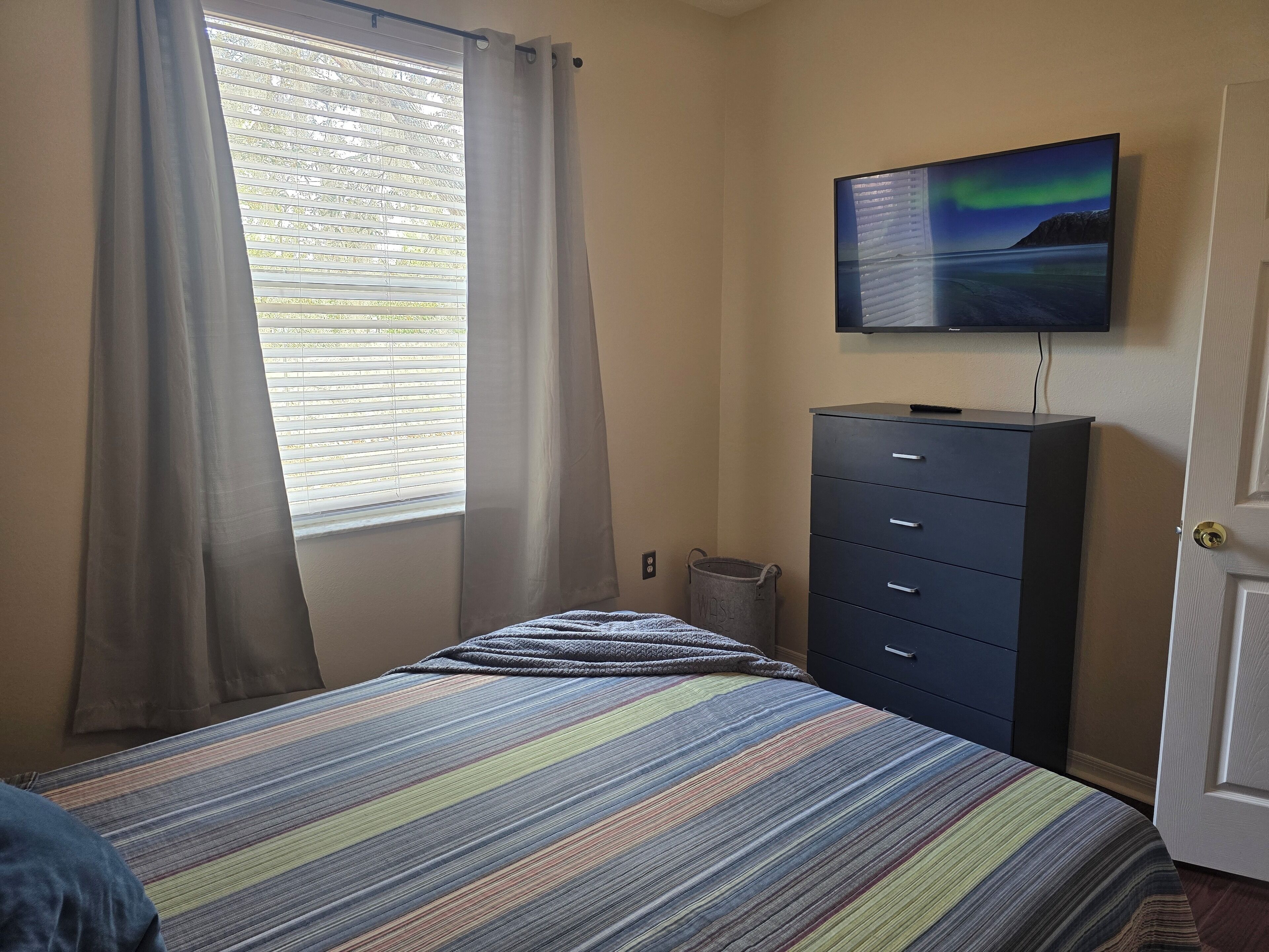 Une chambre à coucher avec un lit, une commode, un téléviseur fixé au mur et une fenêtre avec des rideaux.