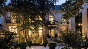 Un edificio luminoso ed elegante, con numerose finestre e balconi, circondato da piante rigogliose e vasi di fiori.