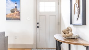 A room with a white door, a framed picture of a lighthouse, a woven basket on a wooden table, and a gray chair.