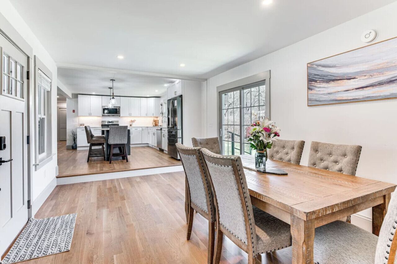 Een moderne keuken met witte kasten, een eethoek met een houten tafel en gestoffeerde stoelen, en een groot raam met uitzicht.