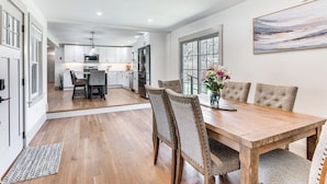 A modern kitchen with white cabinets, a dining area with a wooden table and upholstered chairs, and a large window with a view.