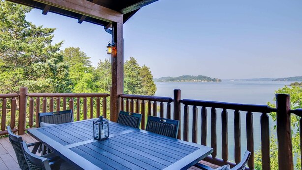 Una terraza de madera con una mesa y sillas, con vista a un lago rodeado de árboles y una orilla lejana.